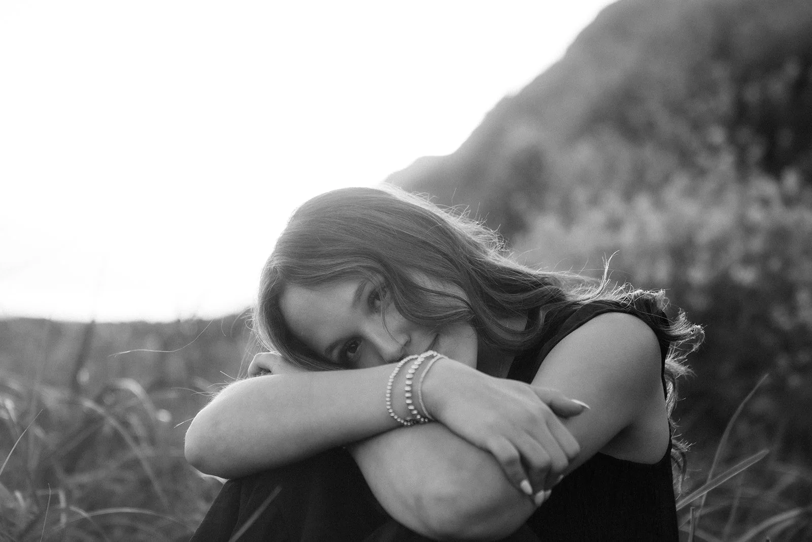 A woman with wavy hair rests her head on her crossed arms outdoors, with a mountain in the background, captured in black and white.