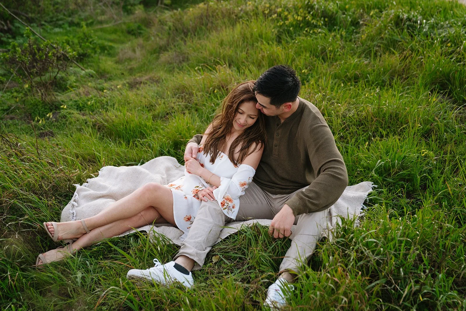 A couple sitting on a blanket in a grassy field, embracing and looking happy.