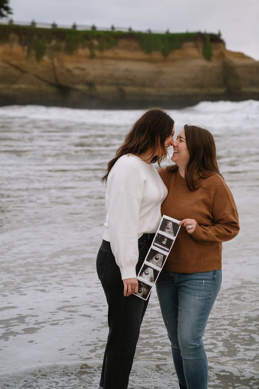Two women stand at the beach, smiling and touching noses, holding a sonogram strip to mark their pregnancy.