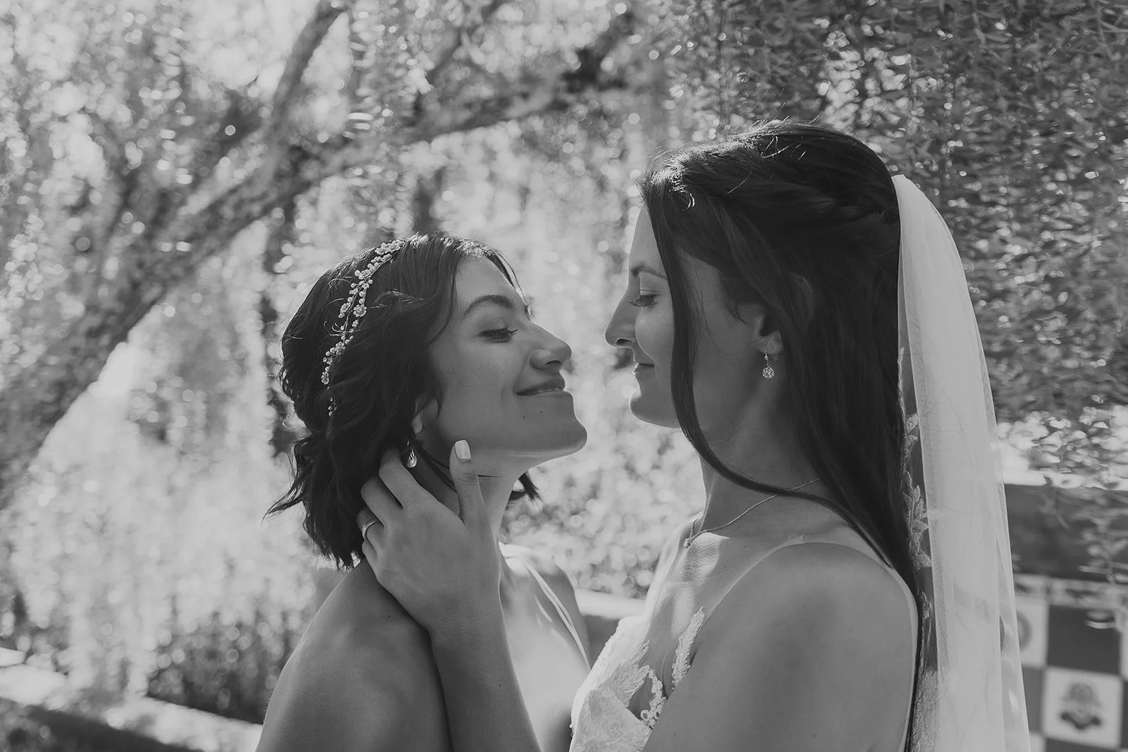 Two women on their wedding day, face-to-face, with one gently holding the other's neck, outdoors with trees in the background, in black and white.