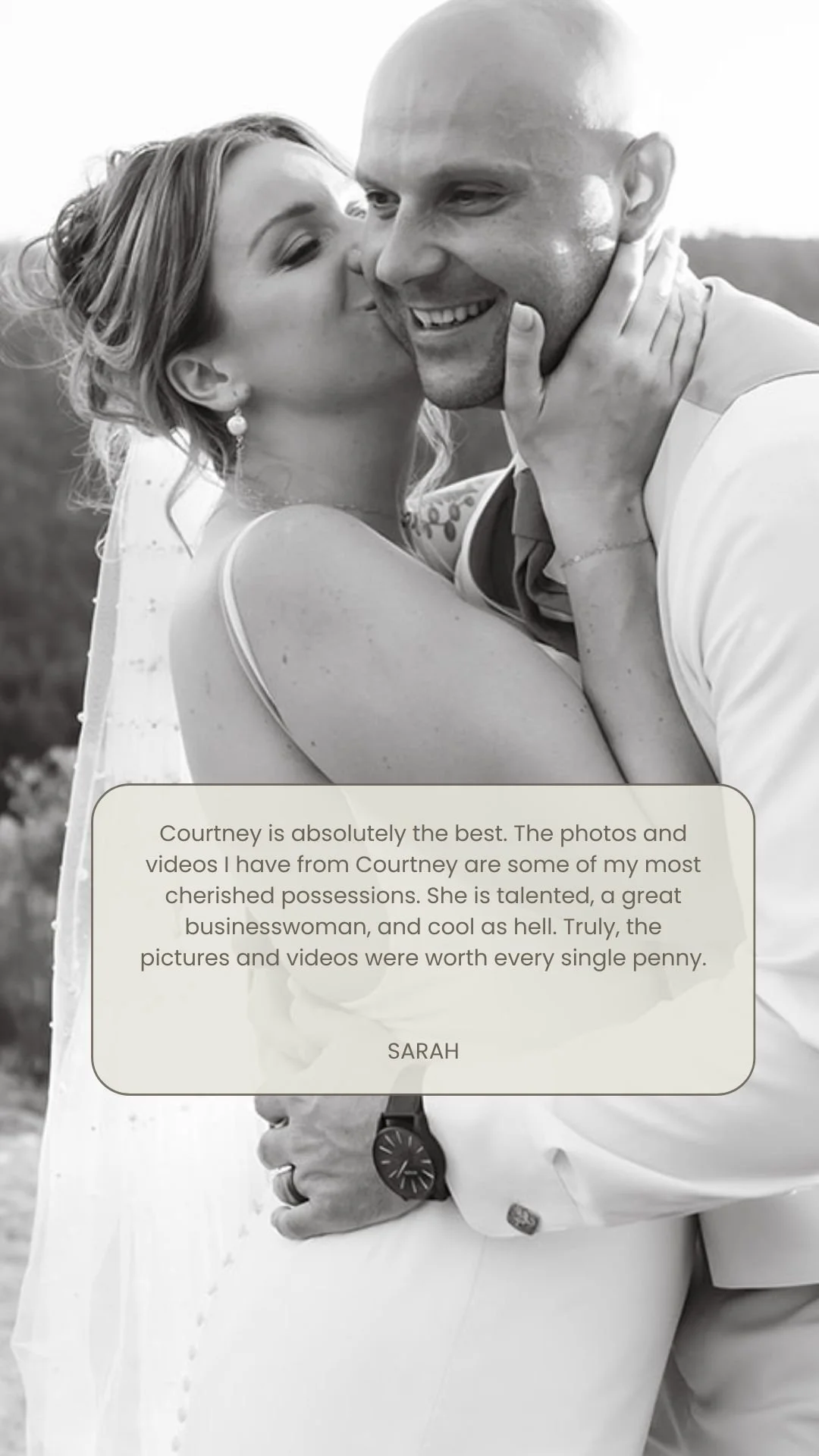 Black and white photo of a woman kissing a man on the cheek. The woman has her hand on the man's face and the man is smiling. The woman is wearing earrings and a dress; the man is in a white shirt.