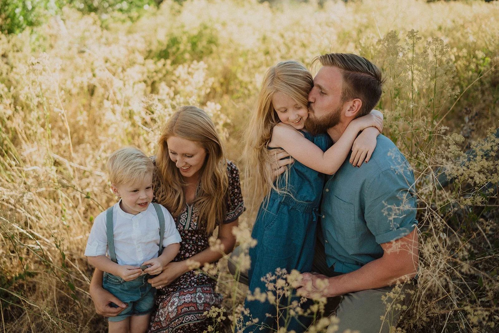 Family of four enjoying time outdoors in a field of tall golden grass, with a woman and man embracing and children sitting nearby.