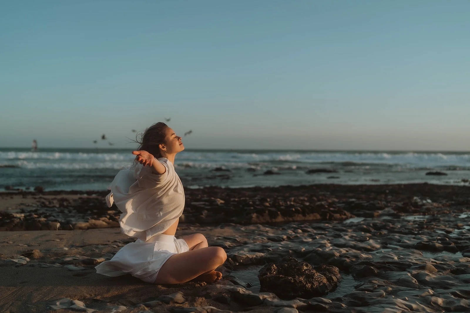 A woman is sitting on a rocky beach with her arms outstretched, enjoying the sunset with her face turned toward the sky, and birds flying in the distance along the ocean horizon.