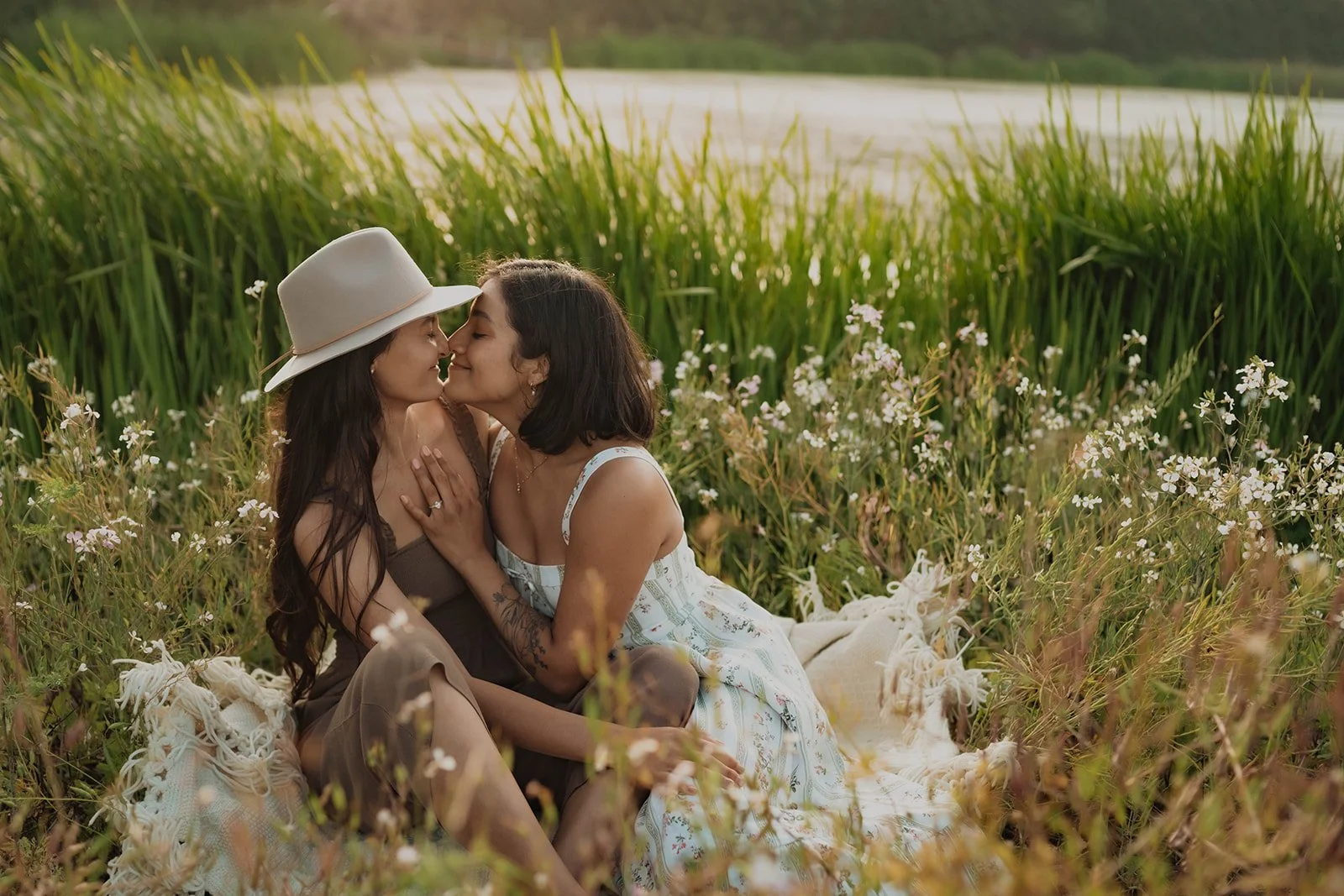 Two women sharing a kiss and smiling, sitting on a blanket in a field of wildflowers near a body of water during sunset