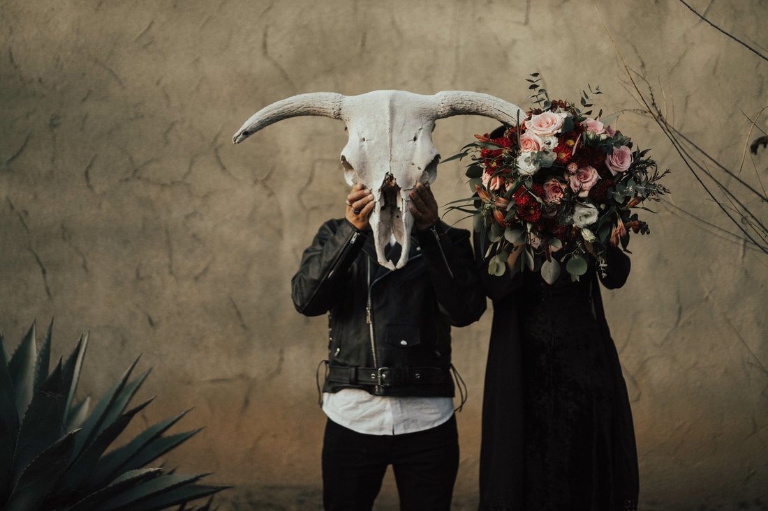 Two people stand side by side, each holding an object in front of their faces. One person holds a large animal skull with horns, and the other holds a bouquet of flowers that covers their face.