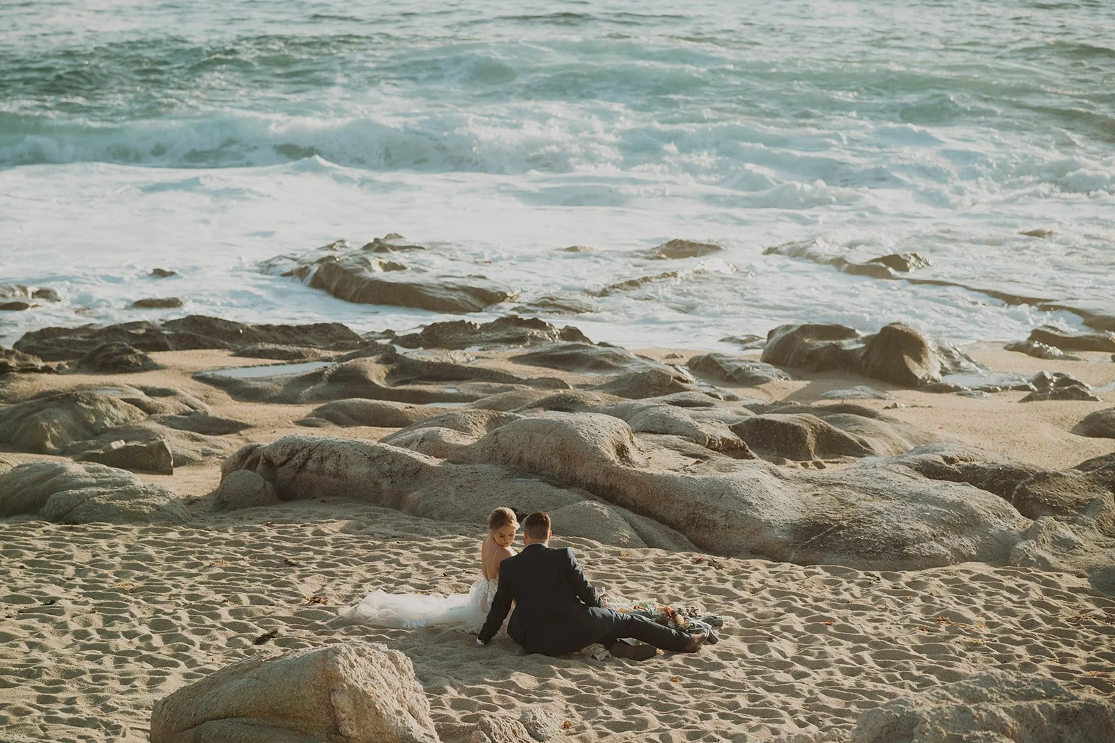 A bride and groom sitting on a sandy beach with rocks, facing the ocean during sunset.