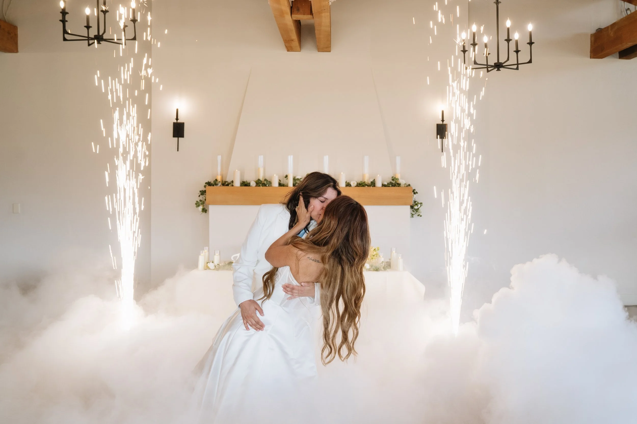 A same-sex couple in wedding attire sharing a kiss during their wedding ceremony, surrounded by smoke and fireworks, inside a decorated venue with candles and chandeliers.