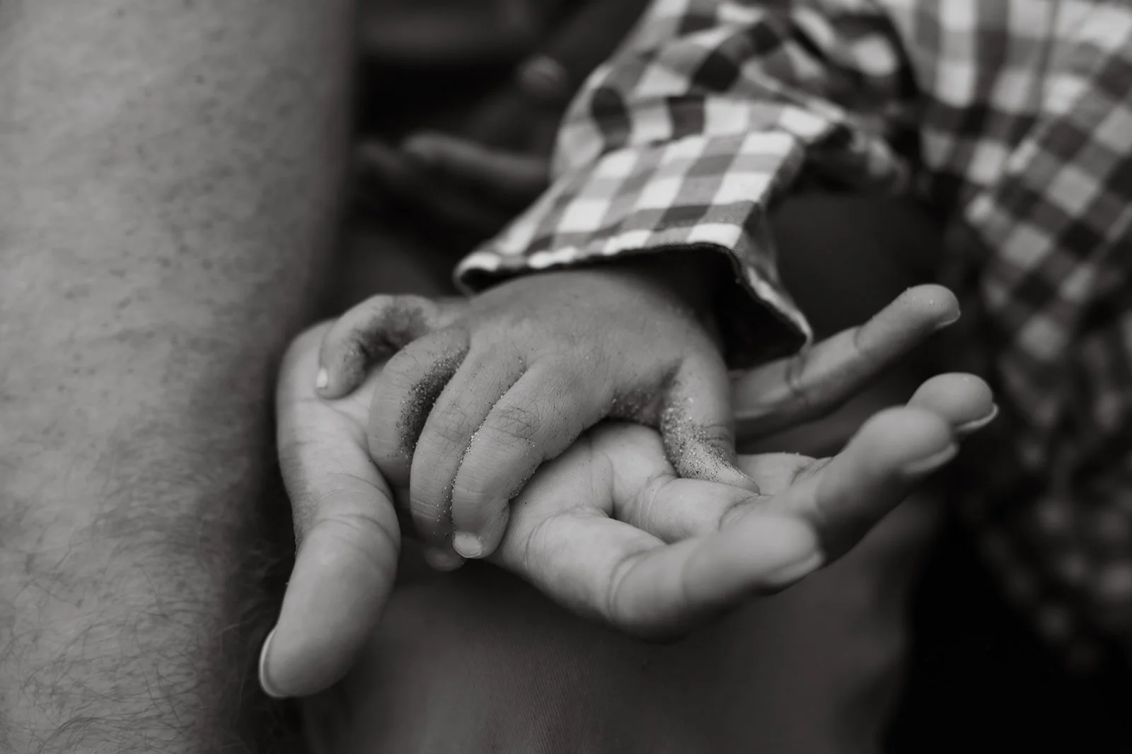 A child with a small, dirty hand holding an adult's finger, belongings on the child. The child is wearing a checkered shirt.