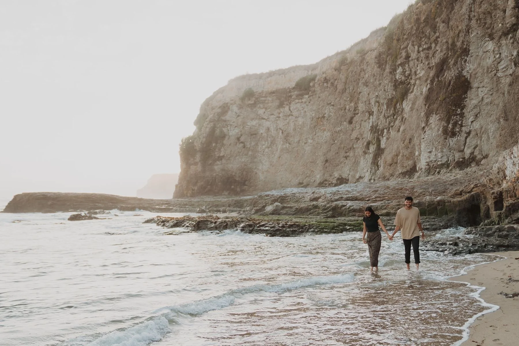 A couple holding hands walking along the shoreline on a beach with cliffs in the background.