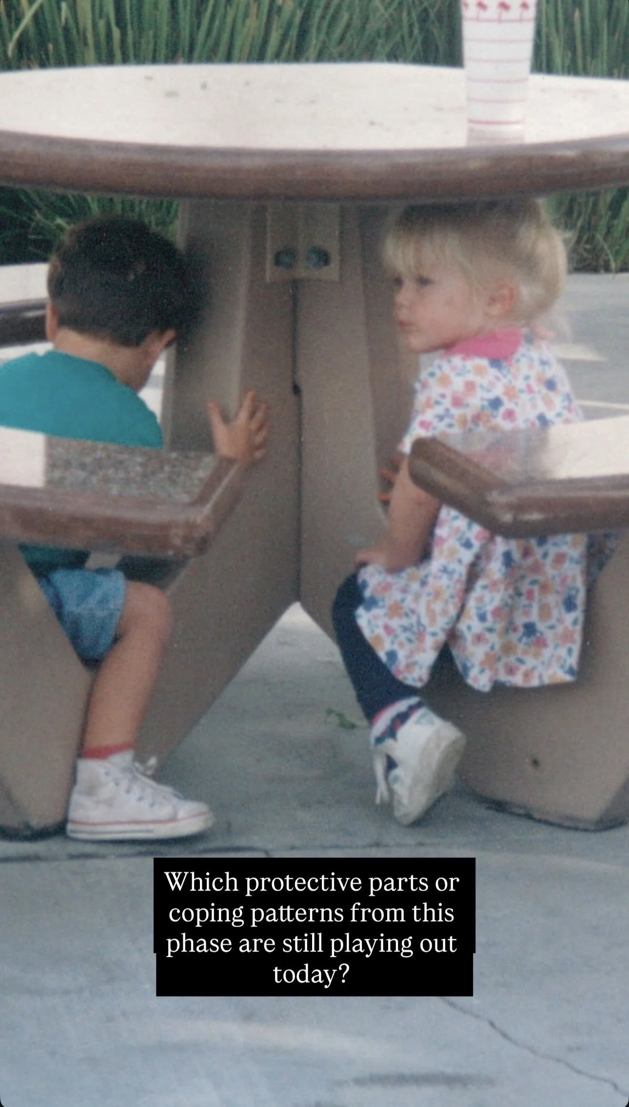 Two children, a boy and a girl, sitting under a picnic table. The boy is wearing a blue shirt and white sneakers, looking towards the corner of the table. The girl, with blonde hair and dressed in a floral dress, looks to the side. There is a caption