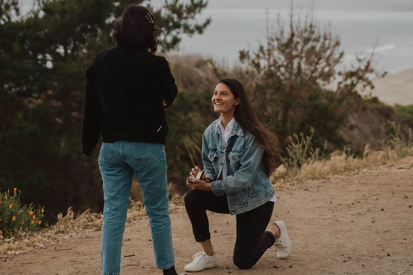 A woman on one knee proposing to another woman on a dirt path with trees and ocean in the background.