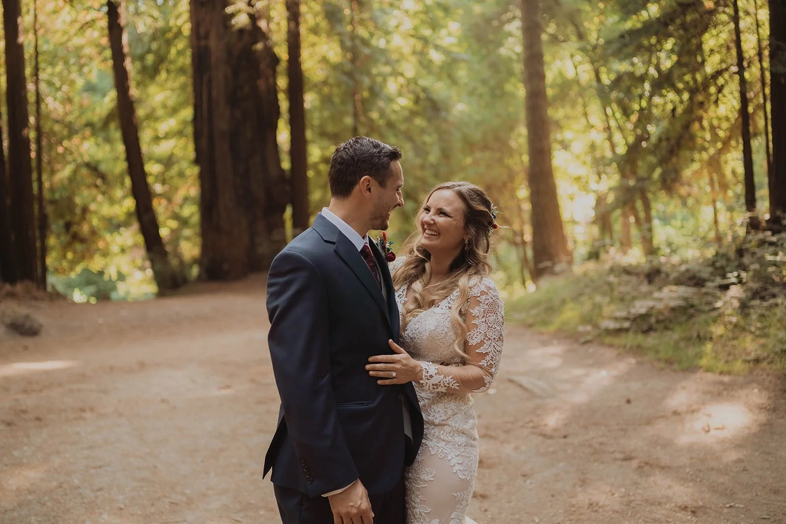 A bride and groom sharing a joyful moment in a forest, dressed in wedding attire, with sunlight filtering through tall trees.