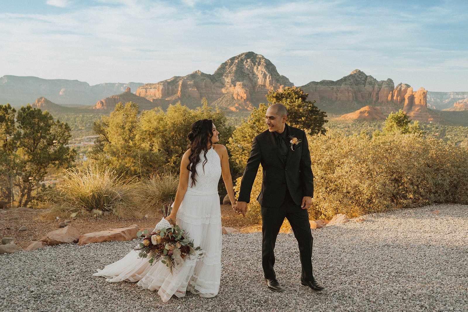A bride and groom holding hands and smiling at each other outdoors with red rock mountains and trees in the background during sunset.