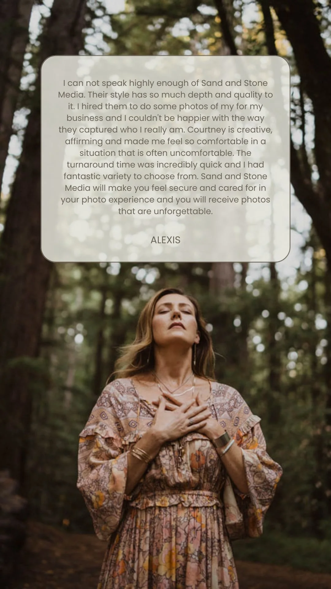 A woman with light brown hair in a patterned dress standing outdoors in a forested area, with her eyes closed and hands over her chest.