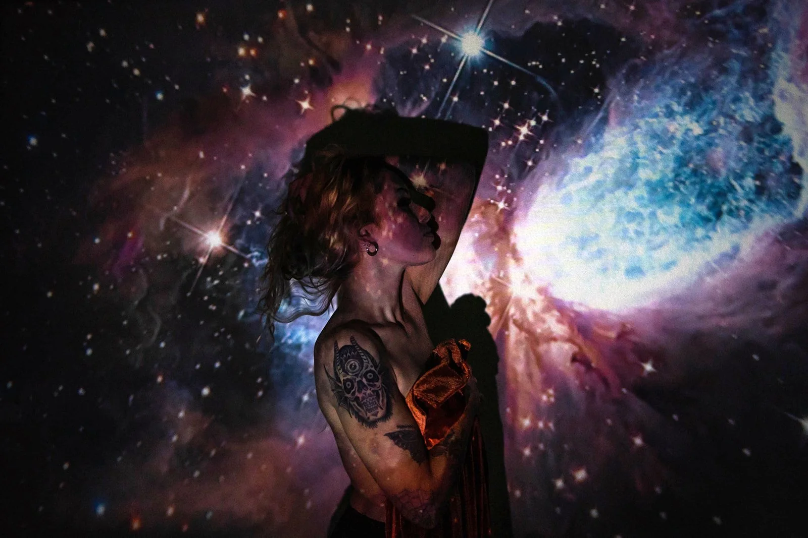 A woman with tattoos on her arm is posing with her arm raised above her head against a backdrop of a colorful galaxy projection, featuring stars, nebulae, and a bright cosmic explosion.
