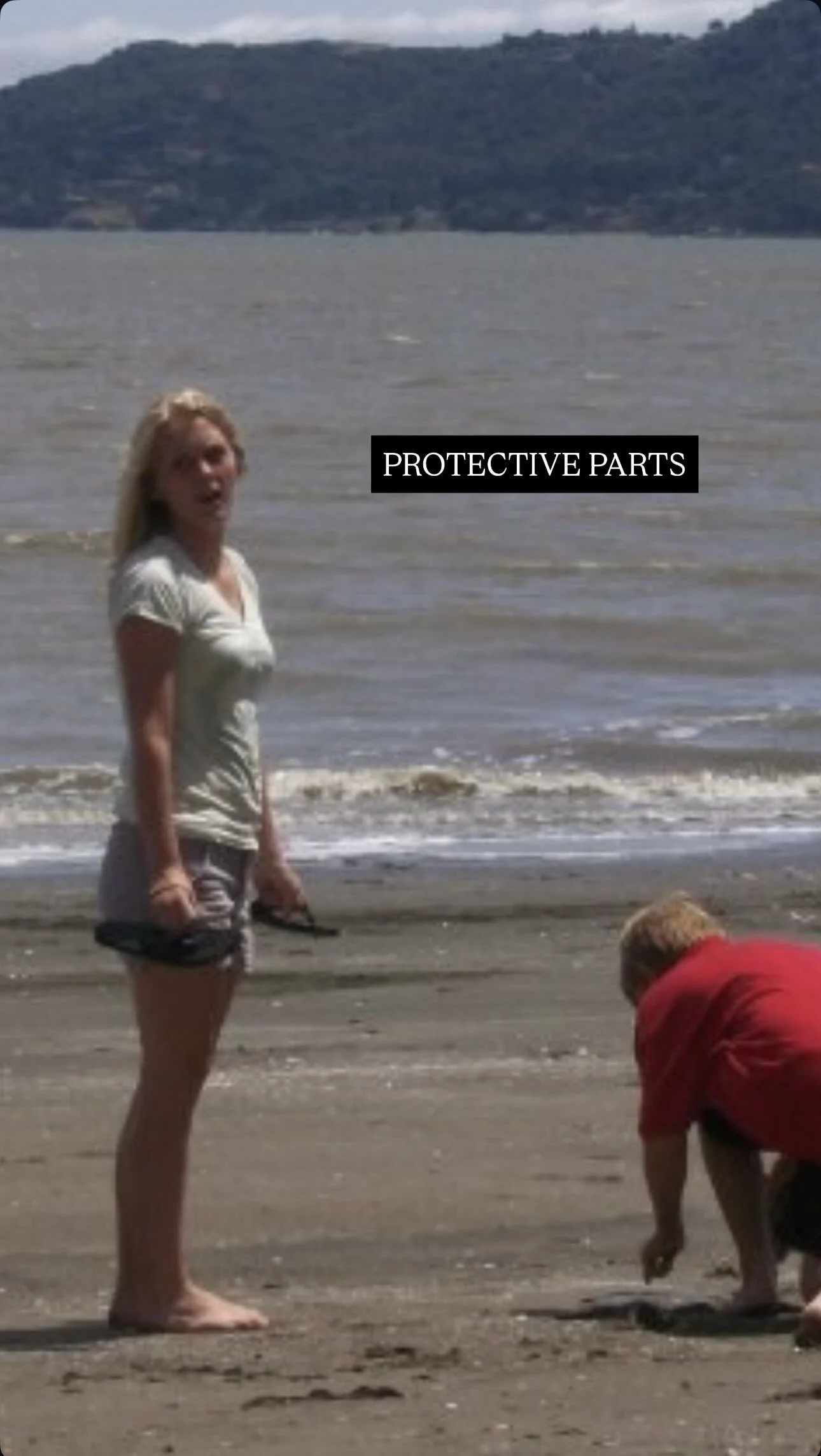 A woman standing on a beach near the water, holding a pair of glasses, with a man crouching down to pick something up. The woman is looking at camera, and there's a label saying 'PROTECTIVE PARTS' across the image.