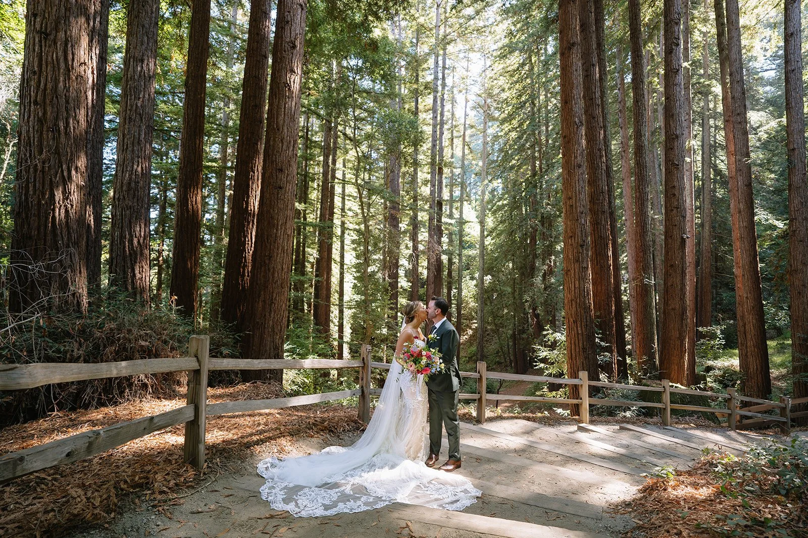 A bride and groom kissing in a forest during their wedding, with tall trees and sunlight filtering through the leaves.