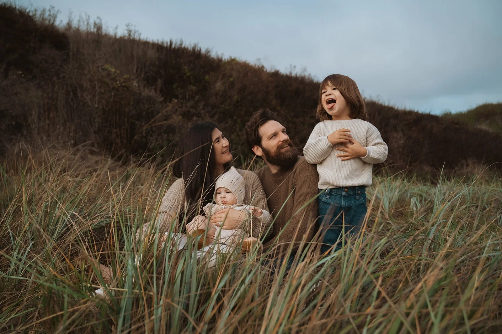 Family of four in a grassy field with hillside in the background, a woman holding a baby, a man holding a child standing, all enjoying a moment of happiness.