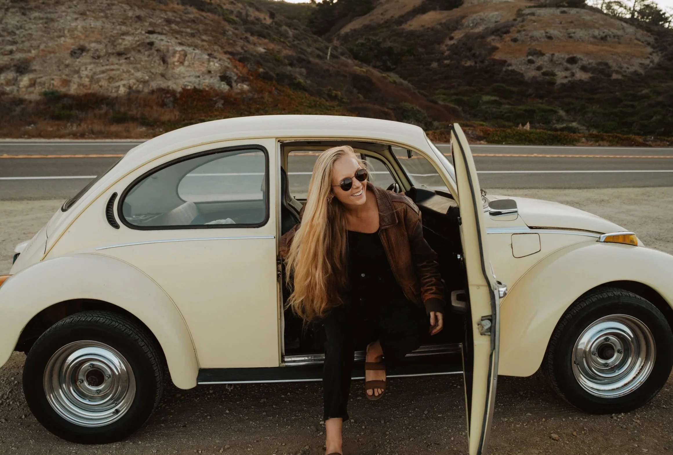A woman with long blonde hair wearing sunglasses, a brown leather jacket, and black pants, smiling as she steps out of a vintage white Volkswagen Beetle parked near a highway with hills in the background.