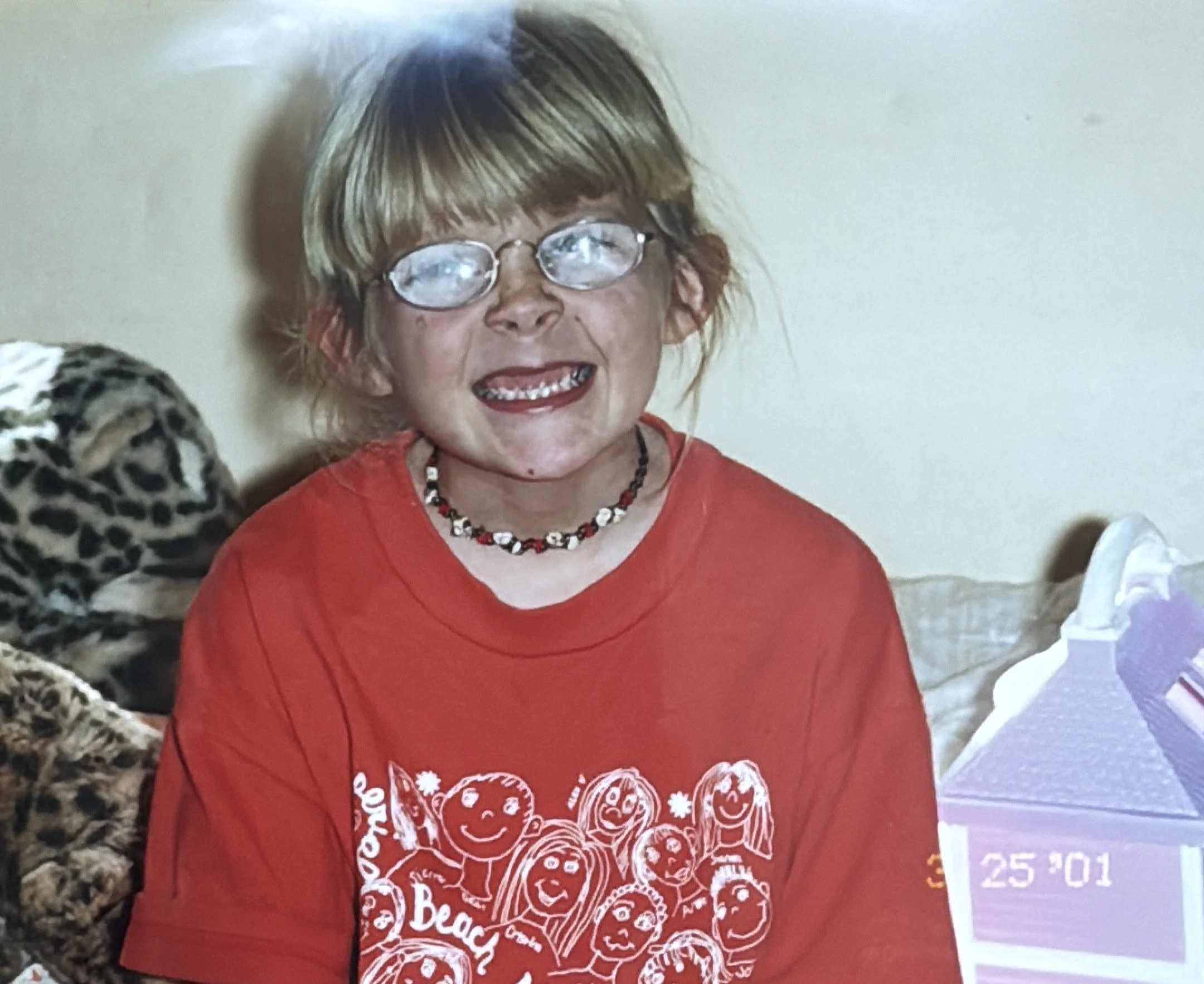 A young girl with glasses and a beaded necklace smiling, wearing a red t-shirt with cartoon drawings of children and text, sitting indoors near a leopard-print blanket or pillow.