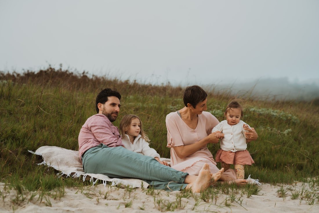 A family of four sitting on a white blanket on the beach overlooking grassy dunes, with a foggy background.