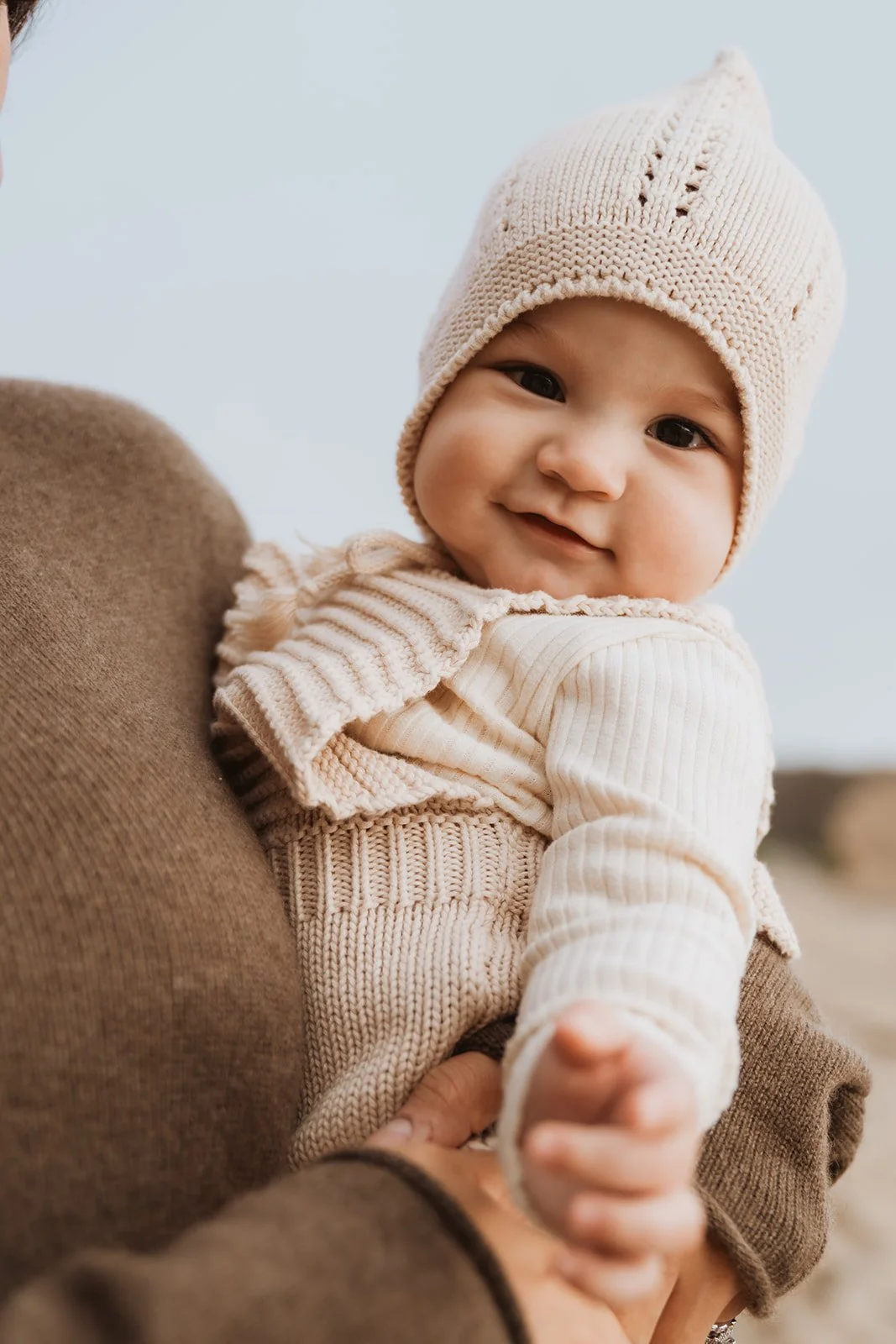 A smiling baby wearing a beige knitted hat and cream-colored sweater, being held outdoors.