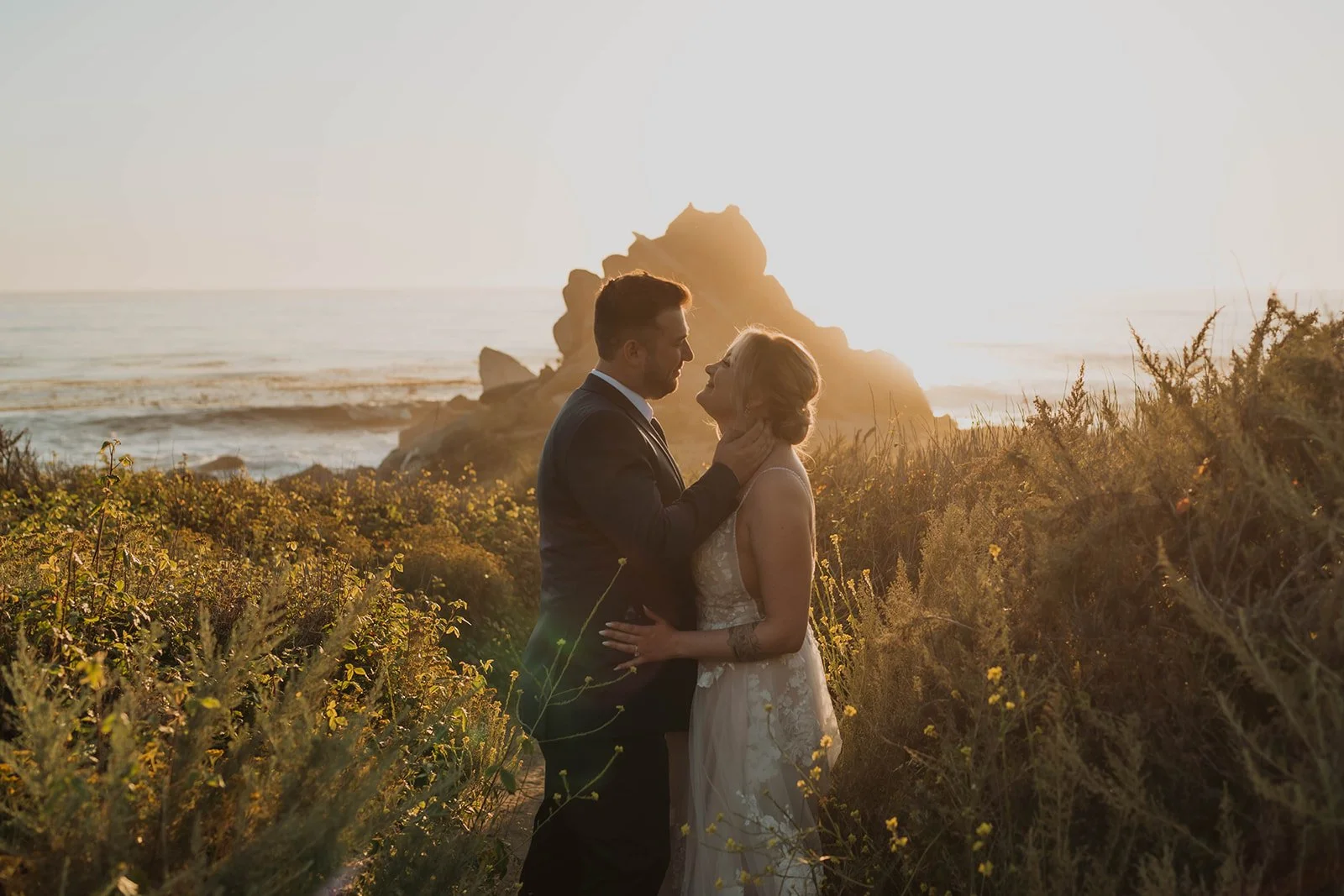 A couple in wedding attire standing close together on a beach at sunset, with waves and rock formations in the background.