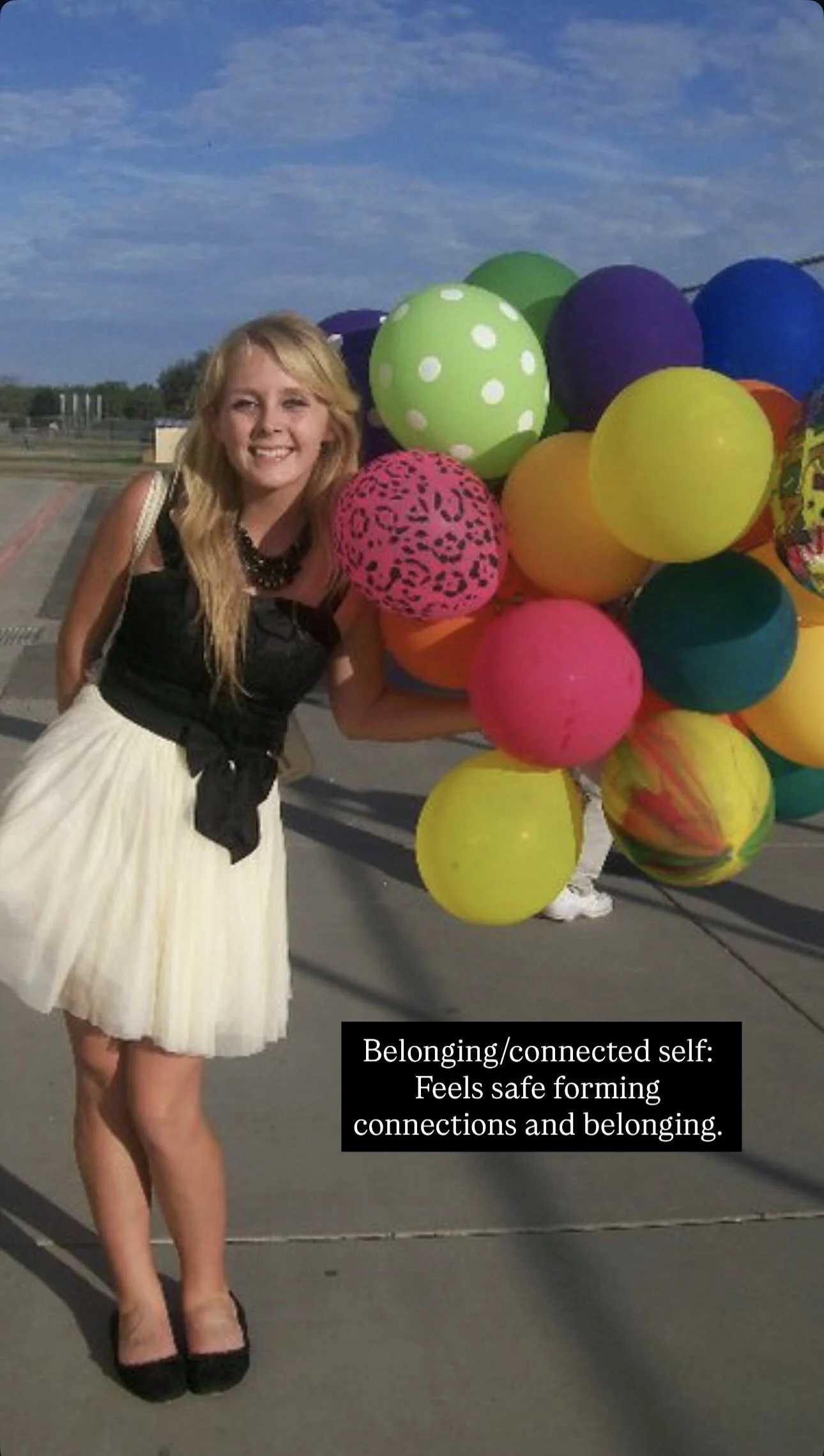 A woman with long blonde hair holding a large bunch of colorful balloons, standing outside on a sunny day, smiling, wearing a black top and white skirt.
