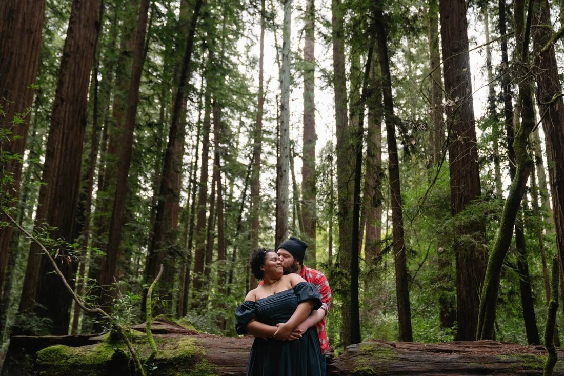 A couple standing close together in a forest, embracing each other while leaning on a fallen tree with tall trees and greenery in the background.