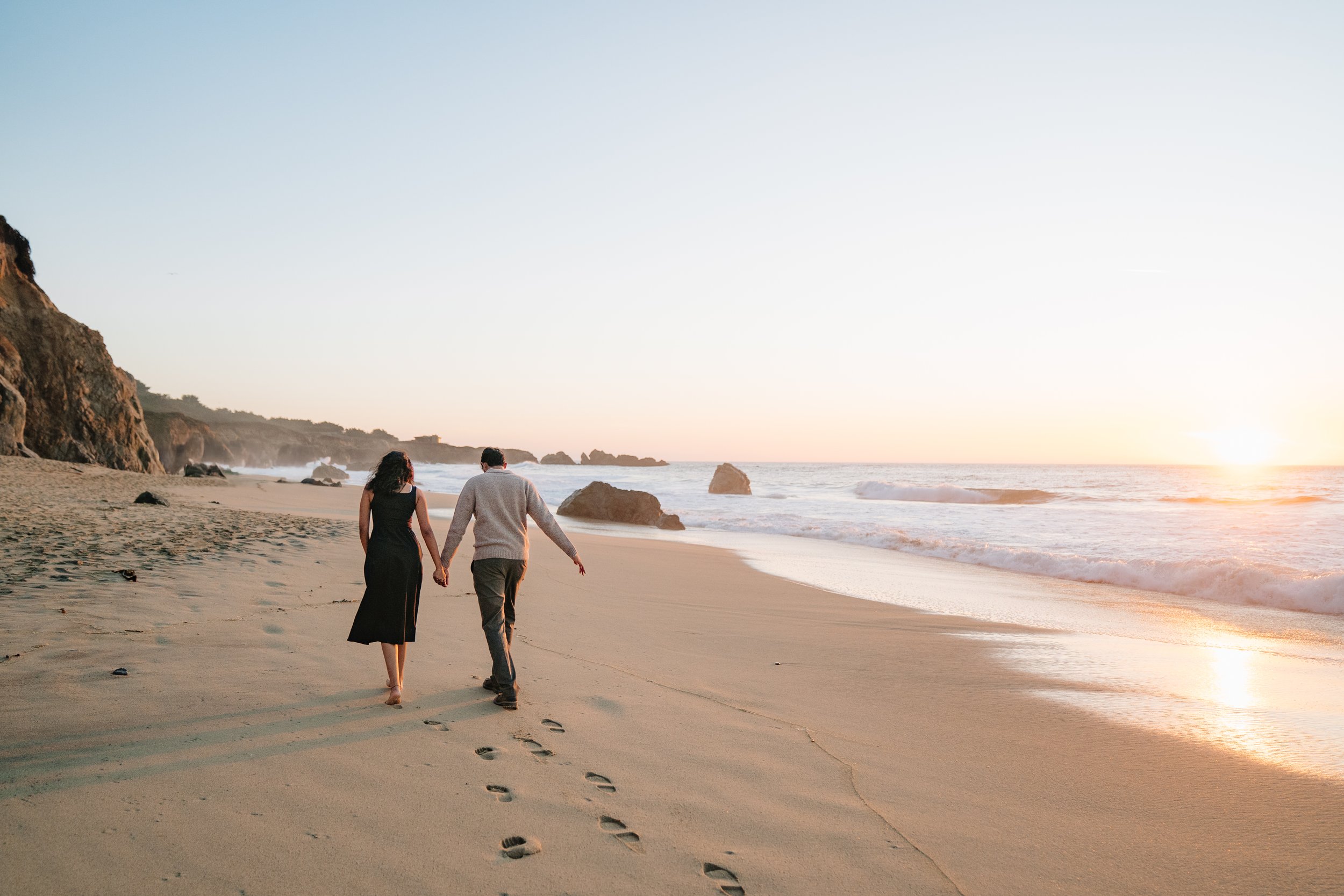 A couple walking hand in hand along the beach at sunset, leaving footprints in the sand with the ocean waves in the background.