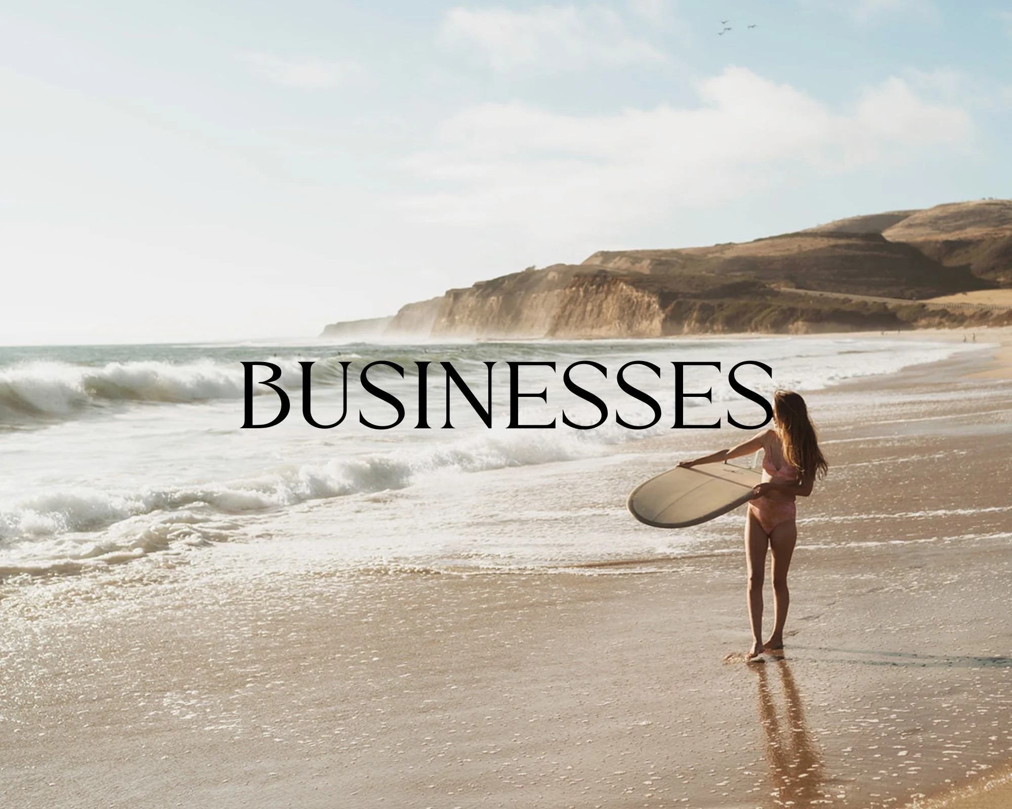 A woman with long hair in a bikini holding a surfboard on a beach facing the ocean with cliffs in the background. The word 'BUSINESSES' is overlaid on the image.