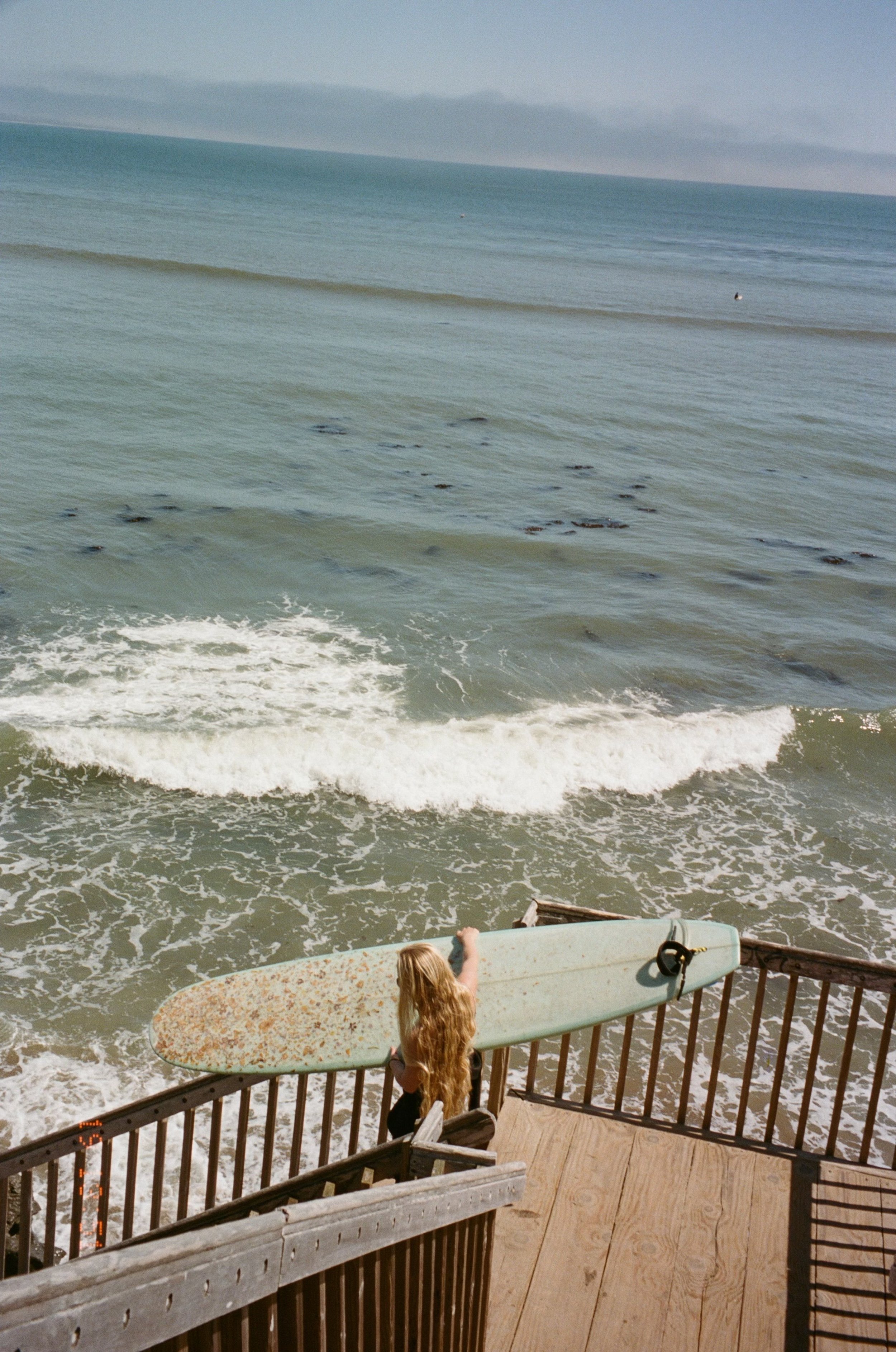 A girl with long blonde hair stands on a wooden deck by the ocean, holding a worn surfboard. The ocean has small waves and a few dark objects in the water, with cloudy skies in the background.