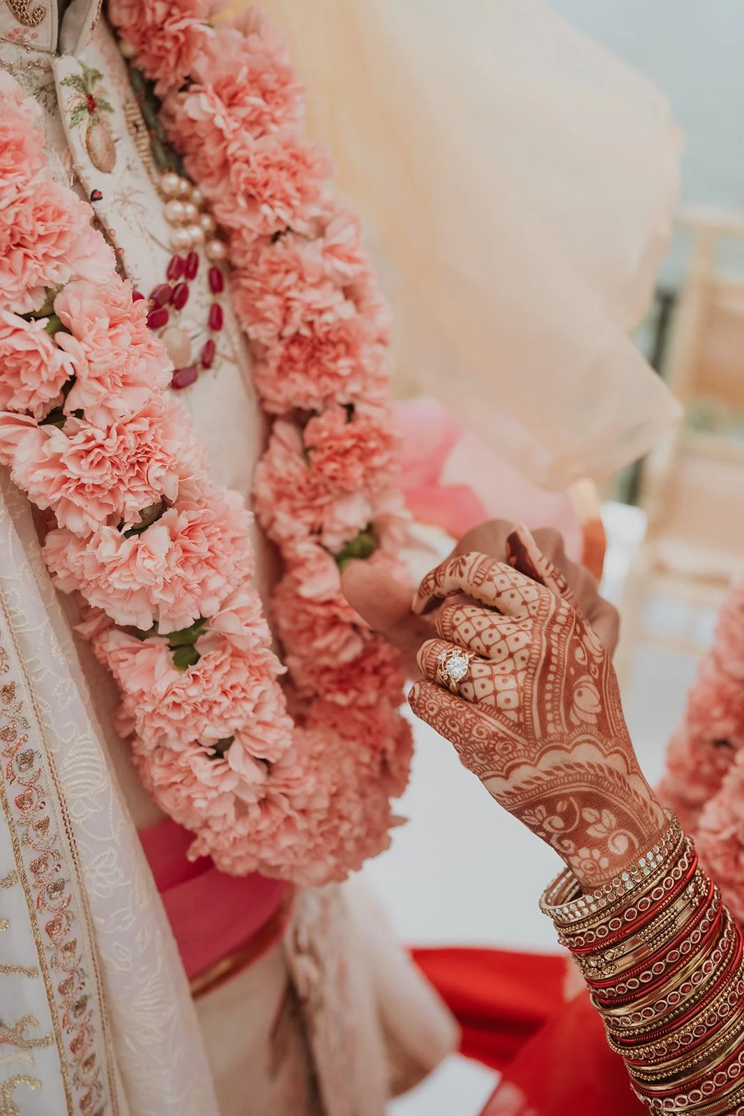 Close-up of a person's hand with intricate henna tattoos and multiple bangles, touching a floral garland on a person dressed in traditional Indian attire with pink flowers, necklaces, and jewelry, during a ceremonial event.
