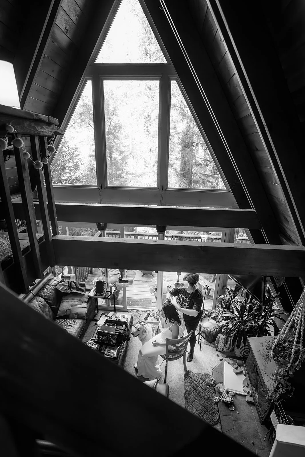 Interior of an A-frame cabin with a high triangular window ceiling. Inside, a woman is sitting in a chair as a hairstylist works on her hair, with a dog seated beside her. The room has various furniture and decorative objects, and natural light streams through the window.