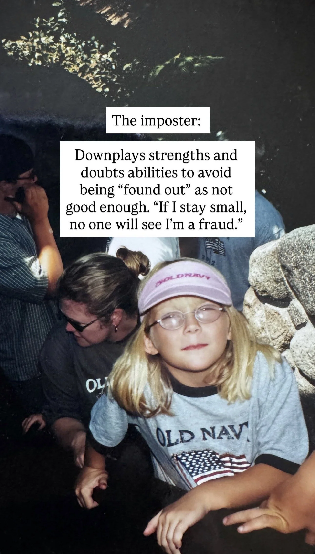 A young girl with blonde hair, glasses, and a pink visor, wearing a gray Old Navy t-shirt with an American flag patch, standing among other people in a dimly lit setting.