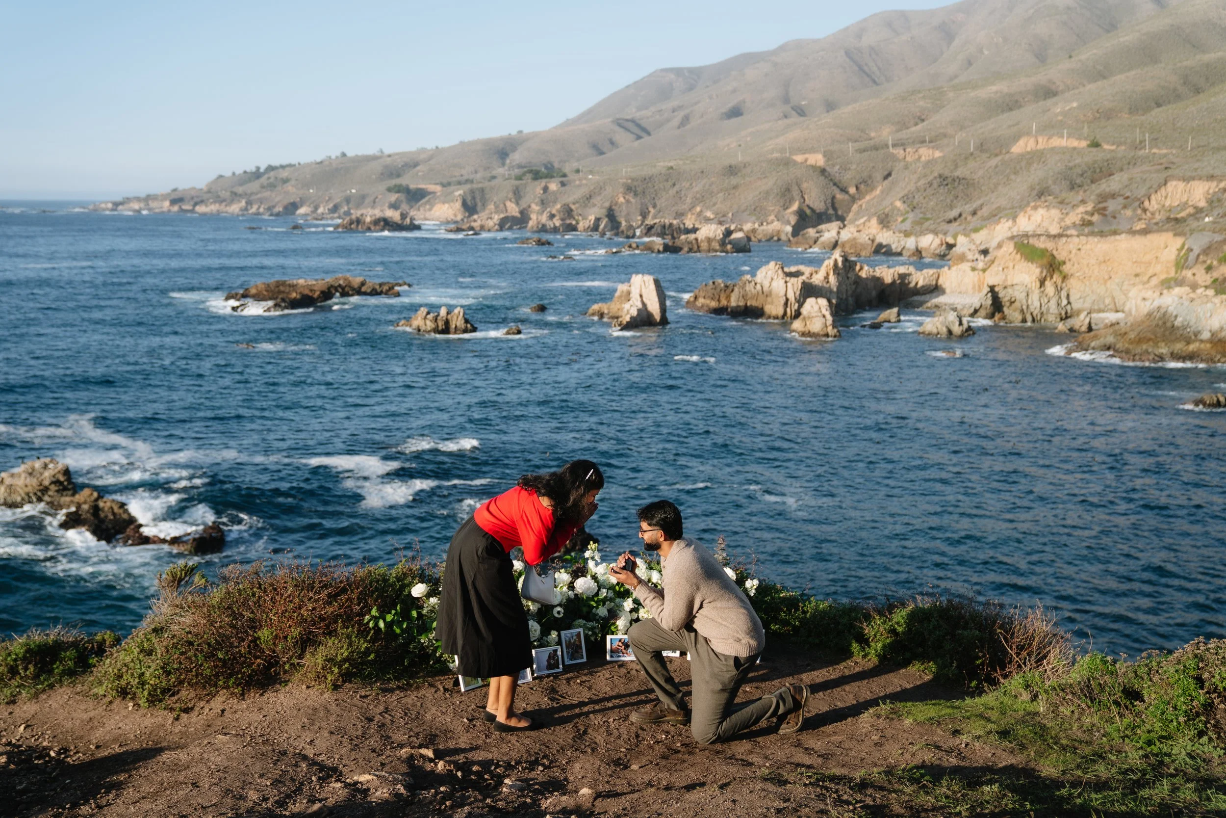 A man proposing marriage to a woman by the seaside with flowers and photos, against a backdrop of rocky coast and hills.