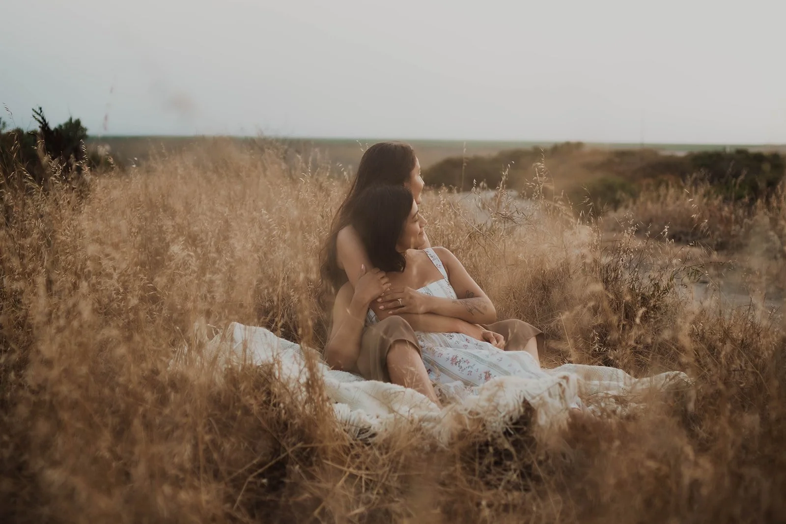 Two women sitting together in a field of tall grass, embracing and looking into the distance on a cloudy day.