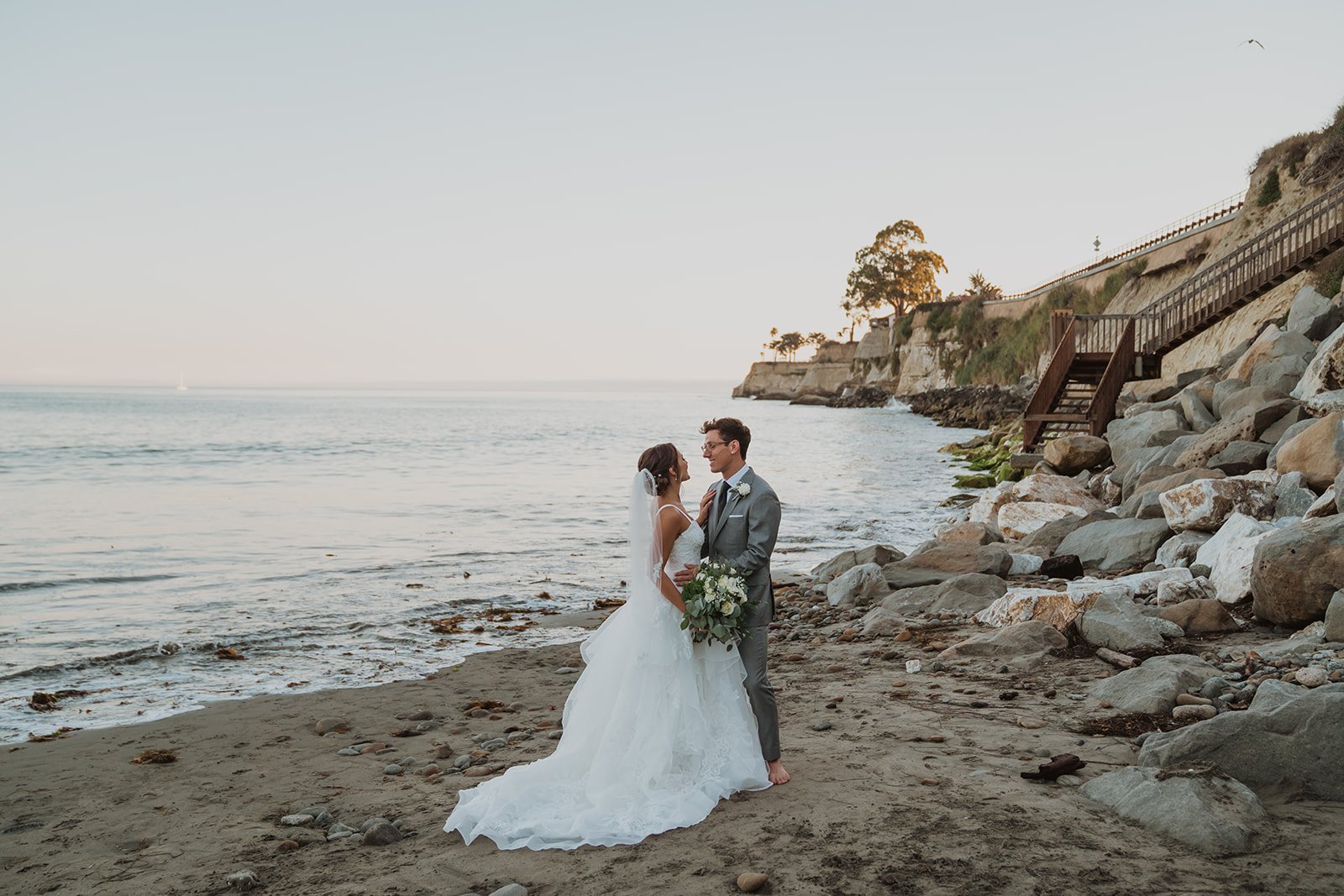 A bride and groom standing barefoot on a beach, facing each other and smiling, with the ocean and rocky shoreline behind them during sunset.