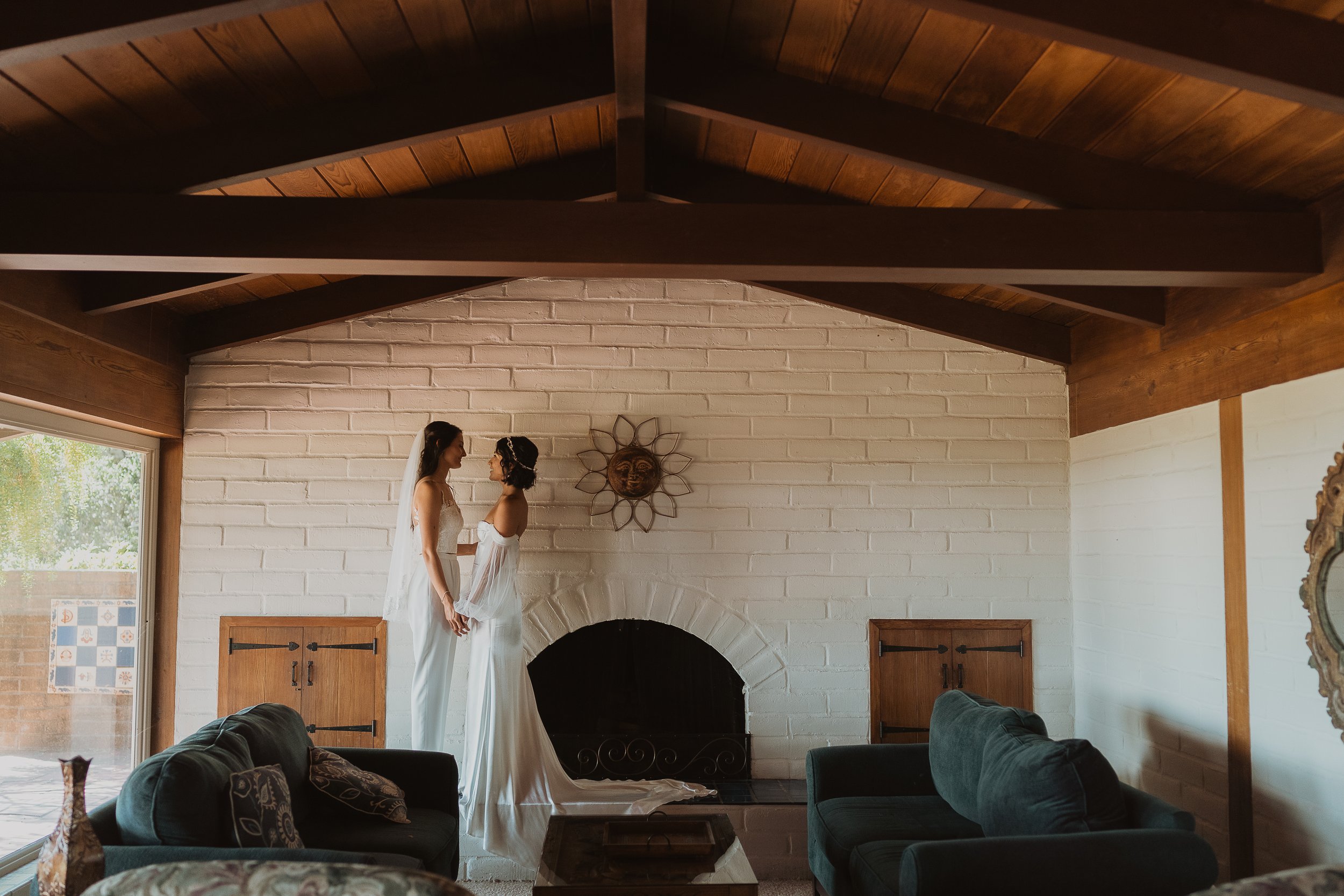 Two women in white dresses standing close to each other in front of a white brick fireplace inside a living room, holding hands and facing each other.