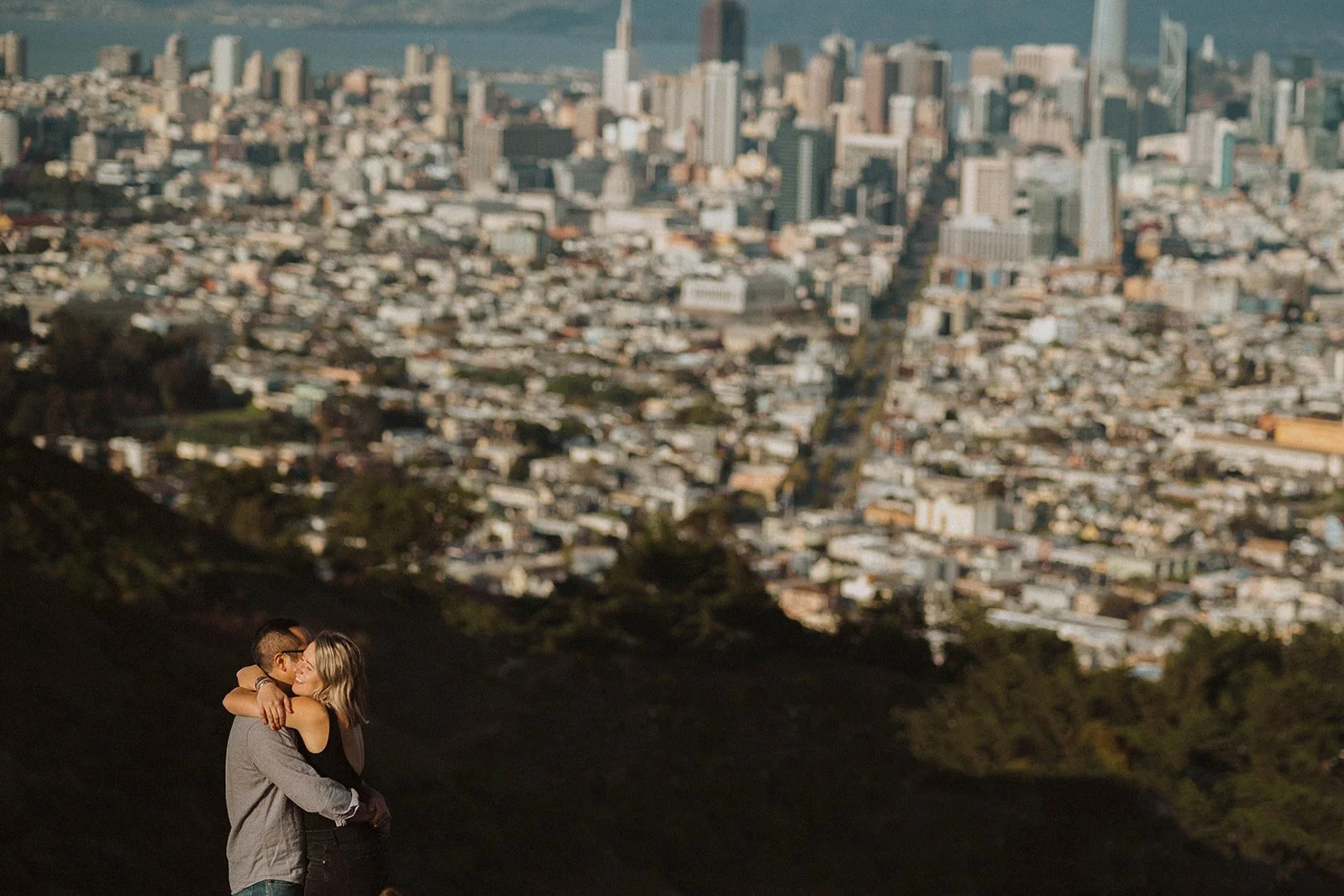 A couple embracing and kissing on a hill overlooking a city skyline with tall buildings.