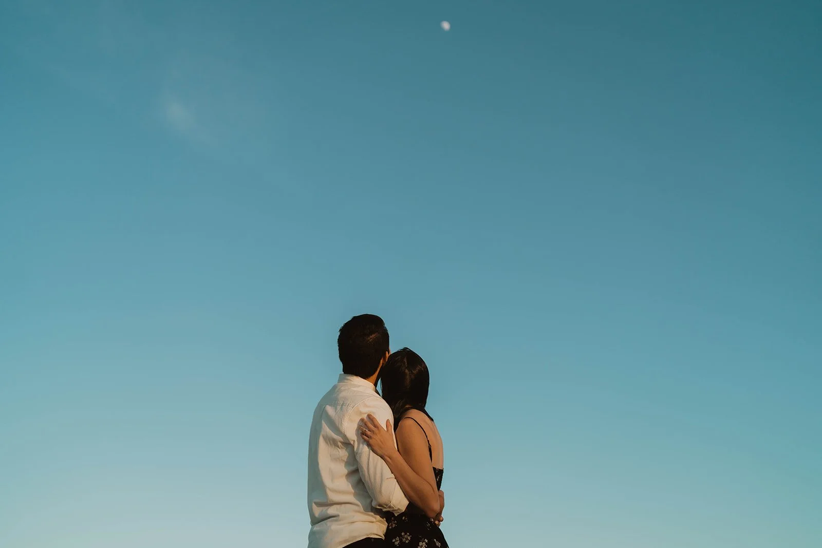 A couple stands close together looking up at the clear blue sky during sunset or twilight hours.