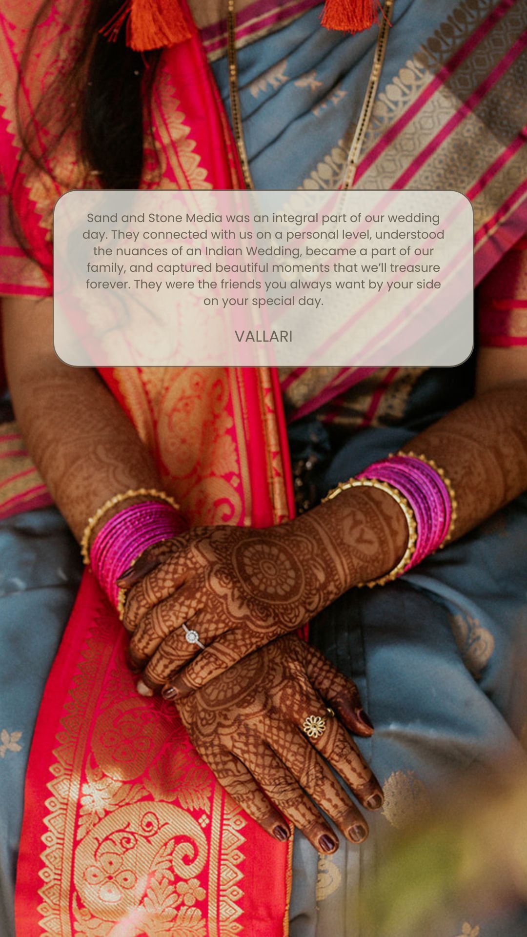 Close-up of a bride's hands with intricate henna designs, wearing rings and bangles, dressed in traditional Indian attire with a red and blue sari, sitting with hands crossed.