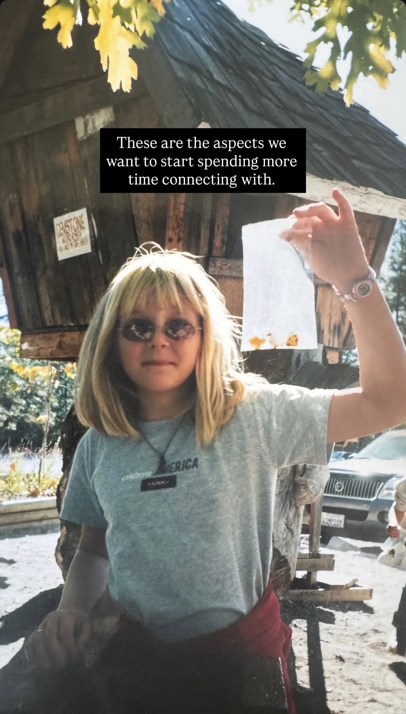 A woman holding a torn paper in front of a wooden structure with trees and cars in the background. She is wearing sunglasses, a gray t-shirt with some text, and a watch, and is making a gesture with her hand.