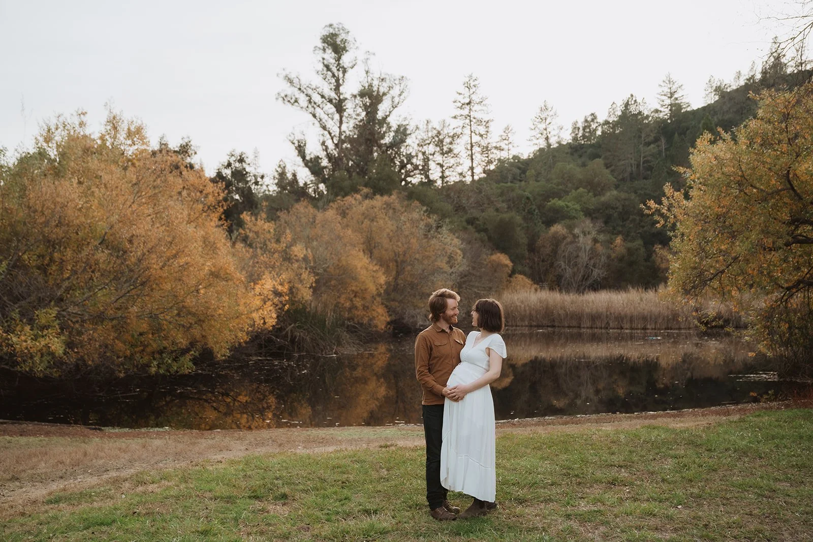 A couple, one pregnant woman in a white dress and a man in a brown shirt, standing close and holding hands near a lake with autumn trees and a hill in the background.