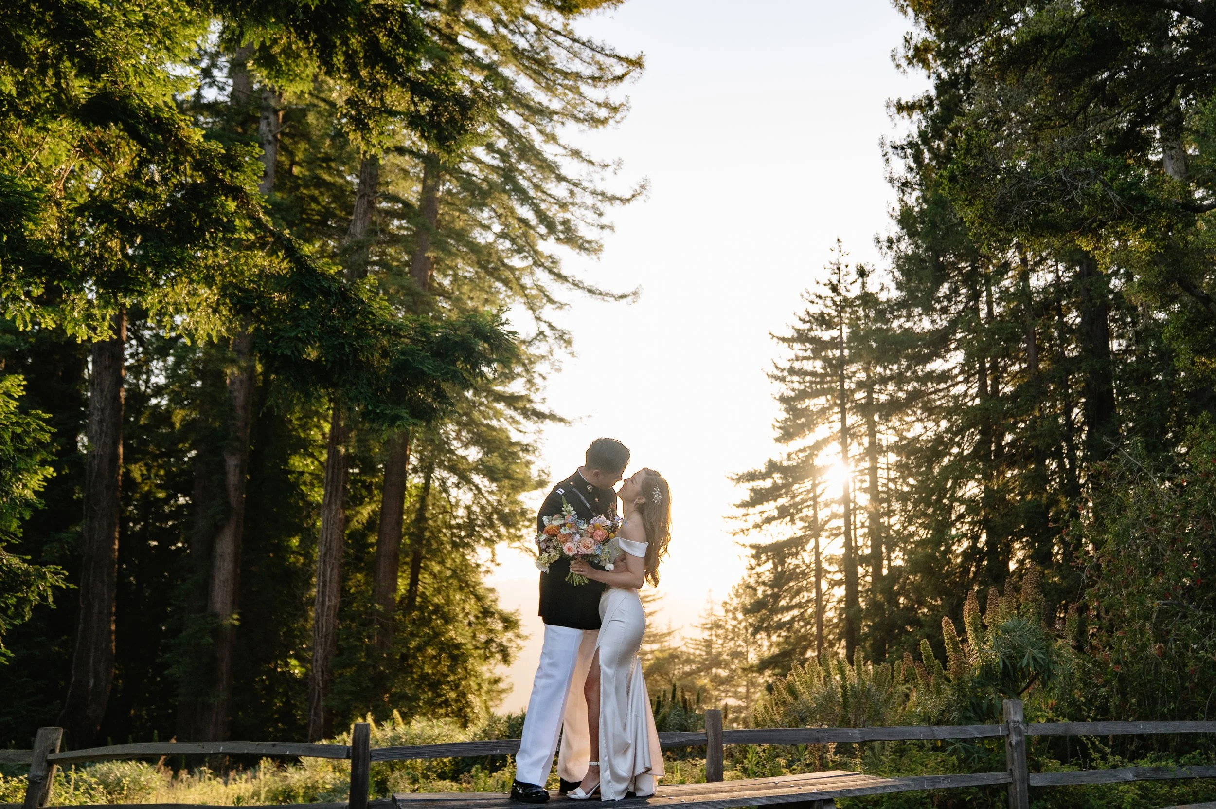 A couple in wedding attire sharing a kiss on a wooden platform surrounded by tall trees during sunset.