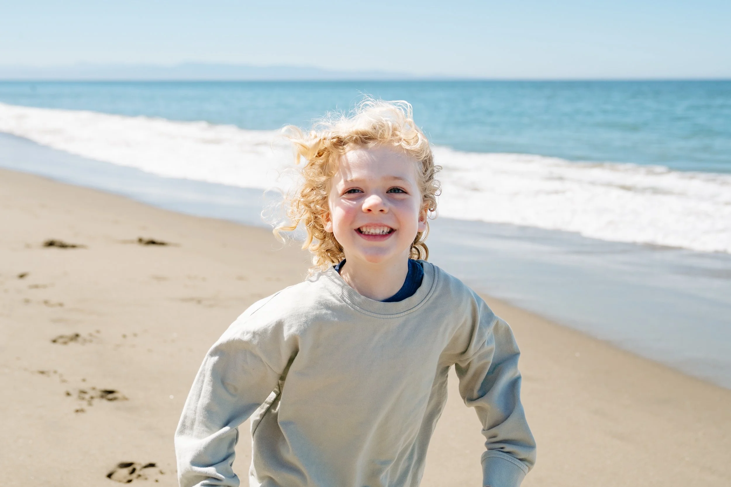 A smiling young boy with curly blonde hair running on a sandy beach, with the ocean and waves in the background.