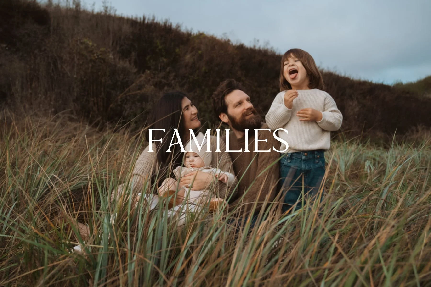 Family of four sitting in tall grass outdoors, with a hillside in the background, enjoying nature together.