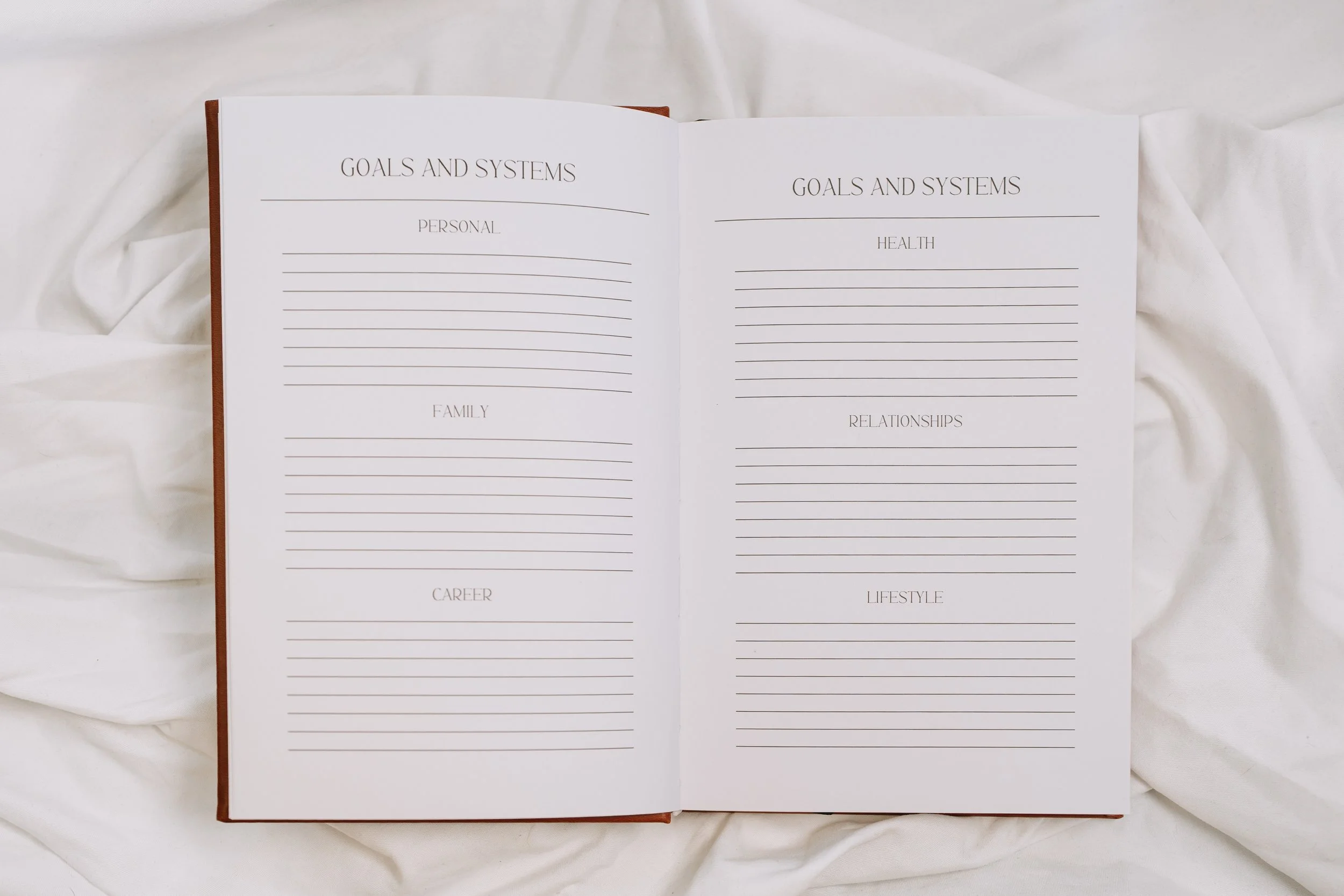An open journal with pages titled 'Goals and Systems' containing sections for personal, family, and career goals on the left page, and health, relationships, and lifestyle goals on the right page, resting on white fabric.