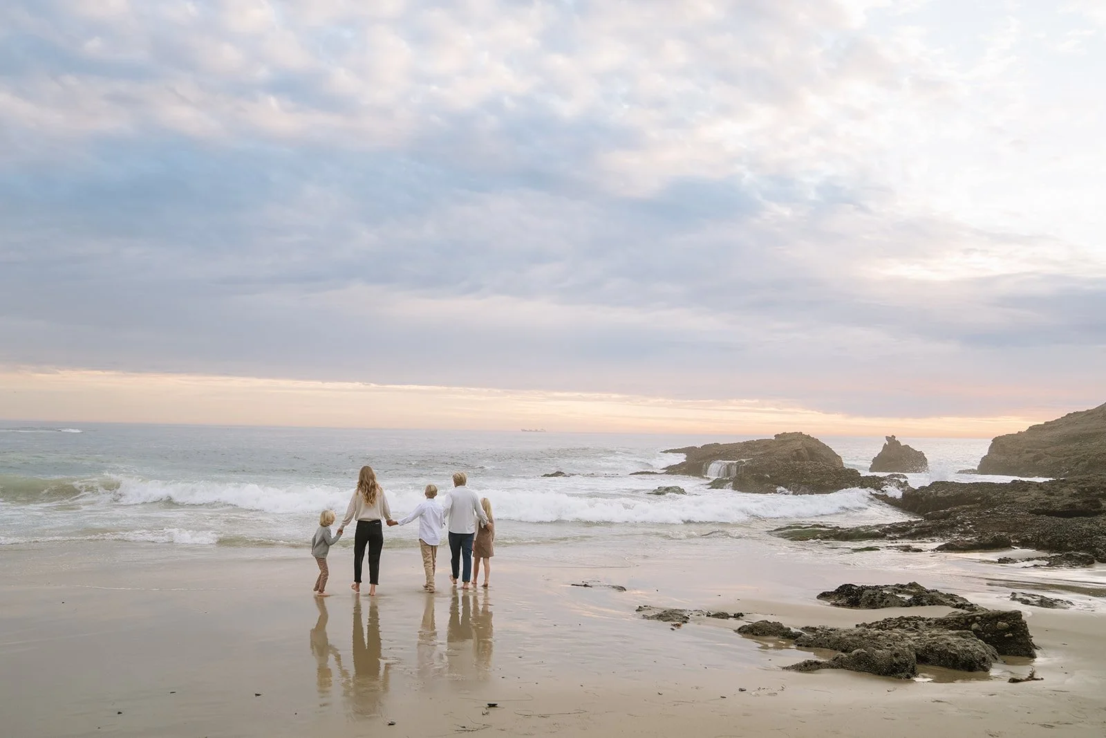 A family of five holding hands and walking along a sandy beach near the ocean at sunset, with rocks and waves in the background.
