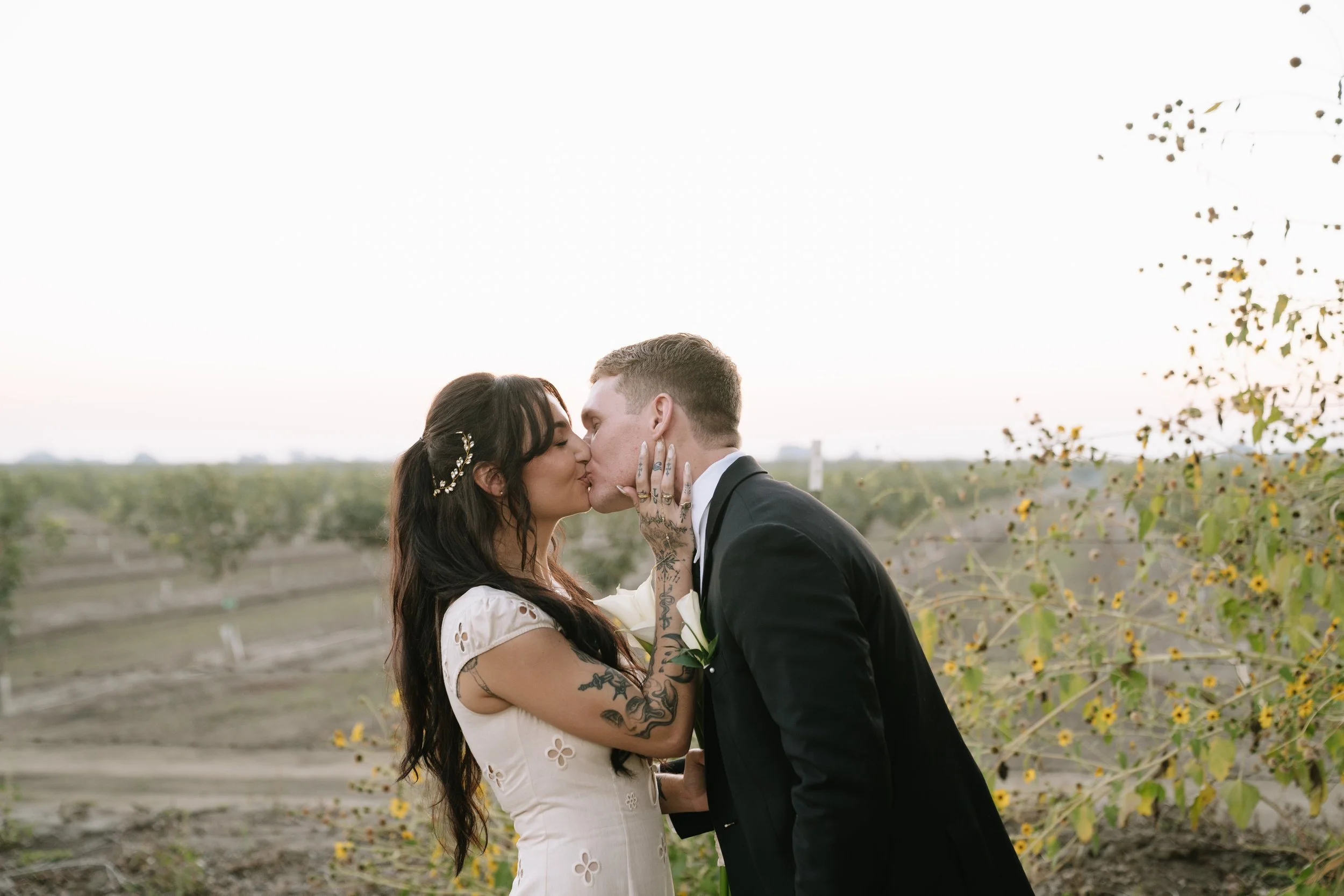 A bride and groom kiss outdoors in a field at sunset